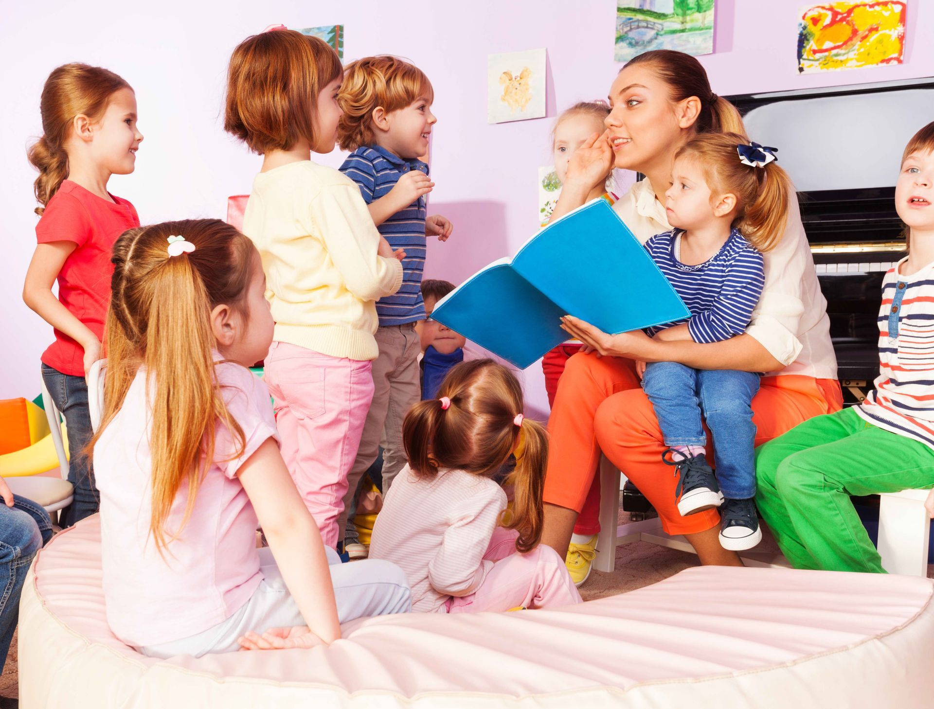 Children sitting and talking together on a bed in a bright playroom, with colorful pillows and a book.