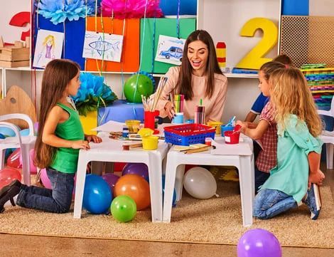 Children at a birthday party table with balloons, colorful gifts, and a smiling adult woman.