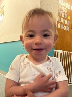 Smiling child in a white shirt indoors, holding one arm across their chest.