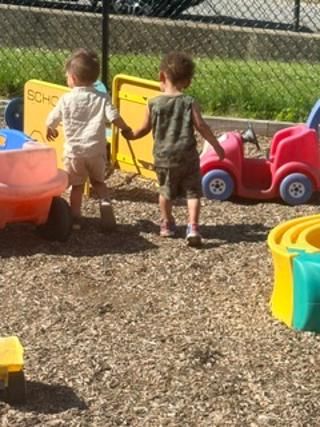 Two children holding hands near colorful toy cars in a fenced outdoor play area