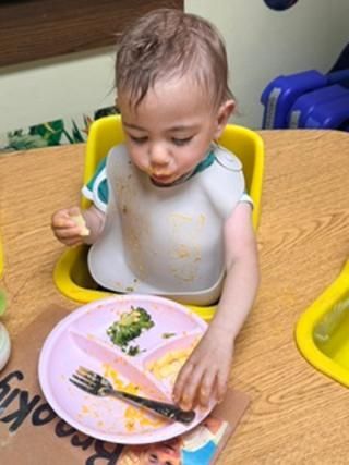 Toddler in a high chair eating from a pink plate at a wooden table.