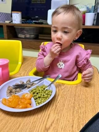 Toddler eating from a plate at a table, with green peas, orange food, and utensils in a yellow high chair.