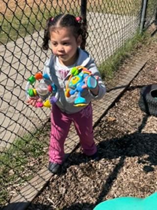 Child holding colorful toys beside a chain-link fence on a sunny path