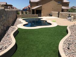 Backyard pool with curved artificial turf and stone landscaping under a bright blue sky