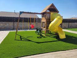 Backyard playground with a yellow spiral slide, swings, and artificial grass under a clear blue sky