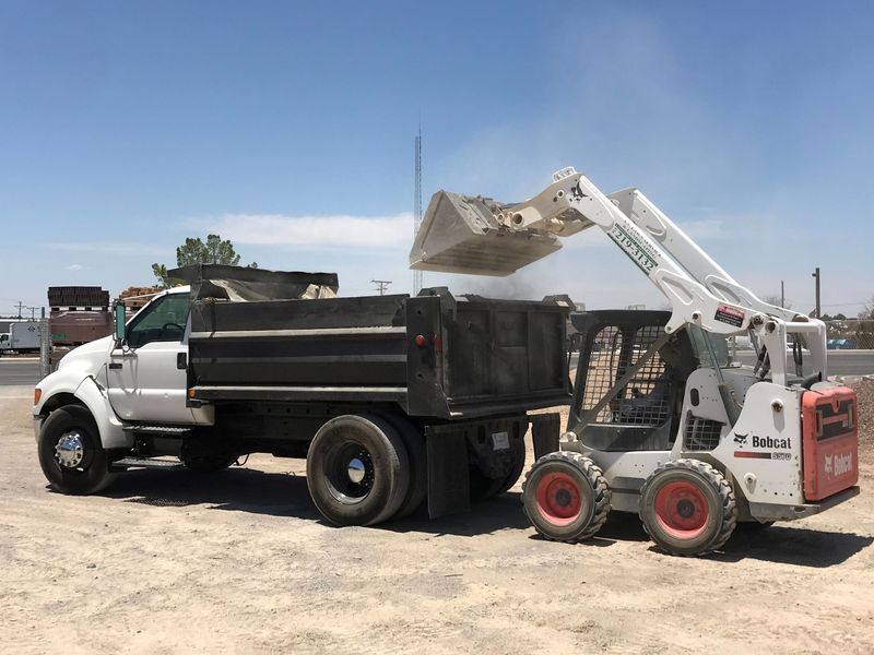 White skid steer loading debris into a black dump truck in a dusty lot under a clear blue sky