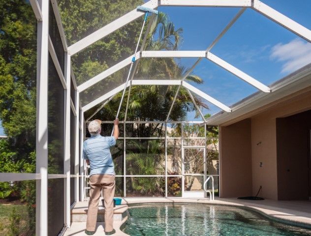 A person in a blue shirt uses a long-handled tool to clean the mesh roof of a residential pool enclosure.
