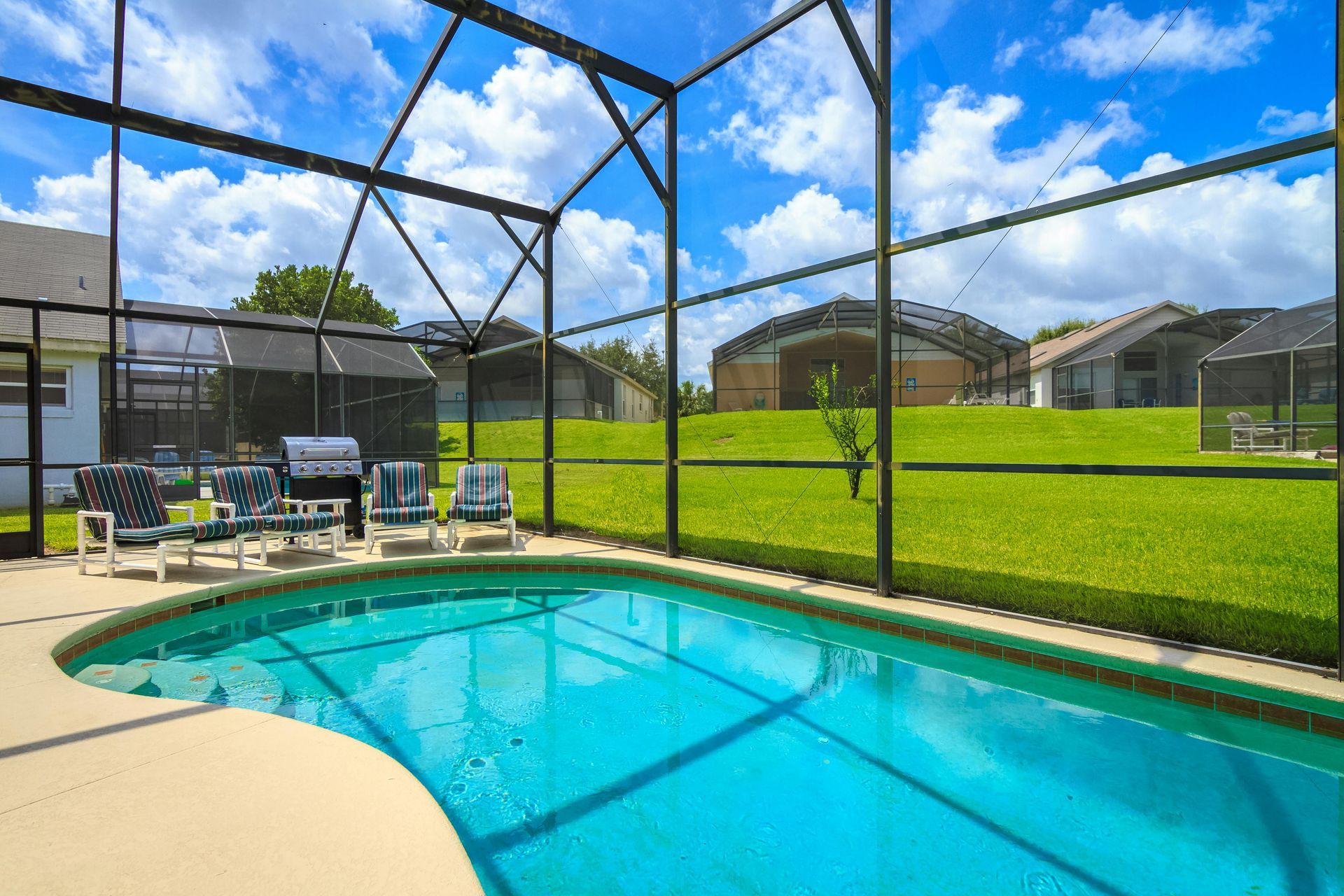 A bright blue swimming pool inside a screened-in lanai, overlooking a grassy backyard under a blue, cloudy sky.