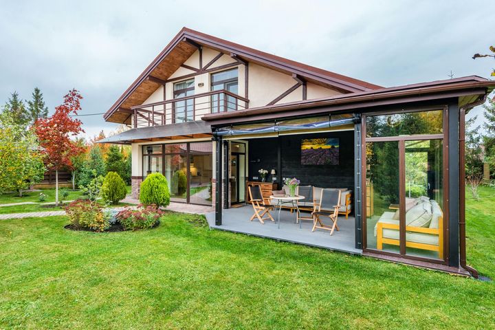 A house with a glass-enclosed patio, wooden furniture, and a balcony set in a lush green backyard on a cloudy day.