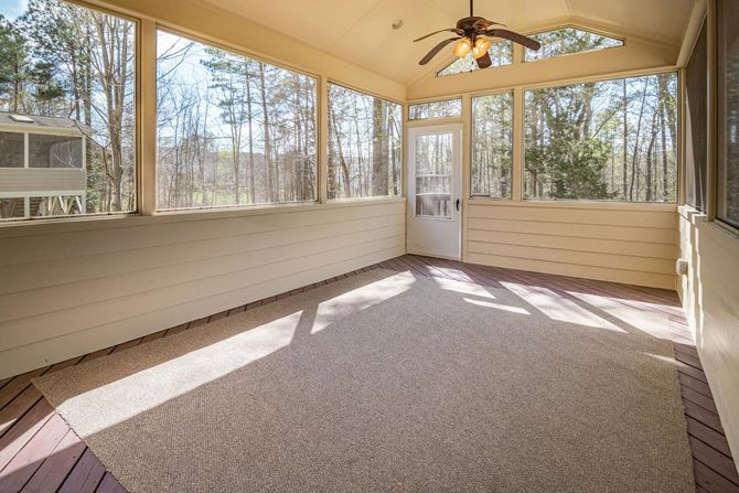 A bright, empty screened-in porch with light-colored wood walls, a large area rug, and a ceiling fan overlooking woods.