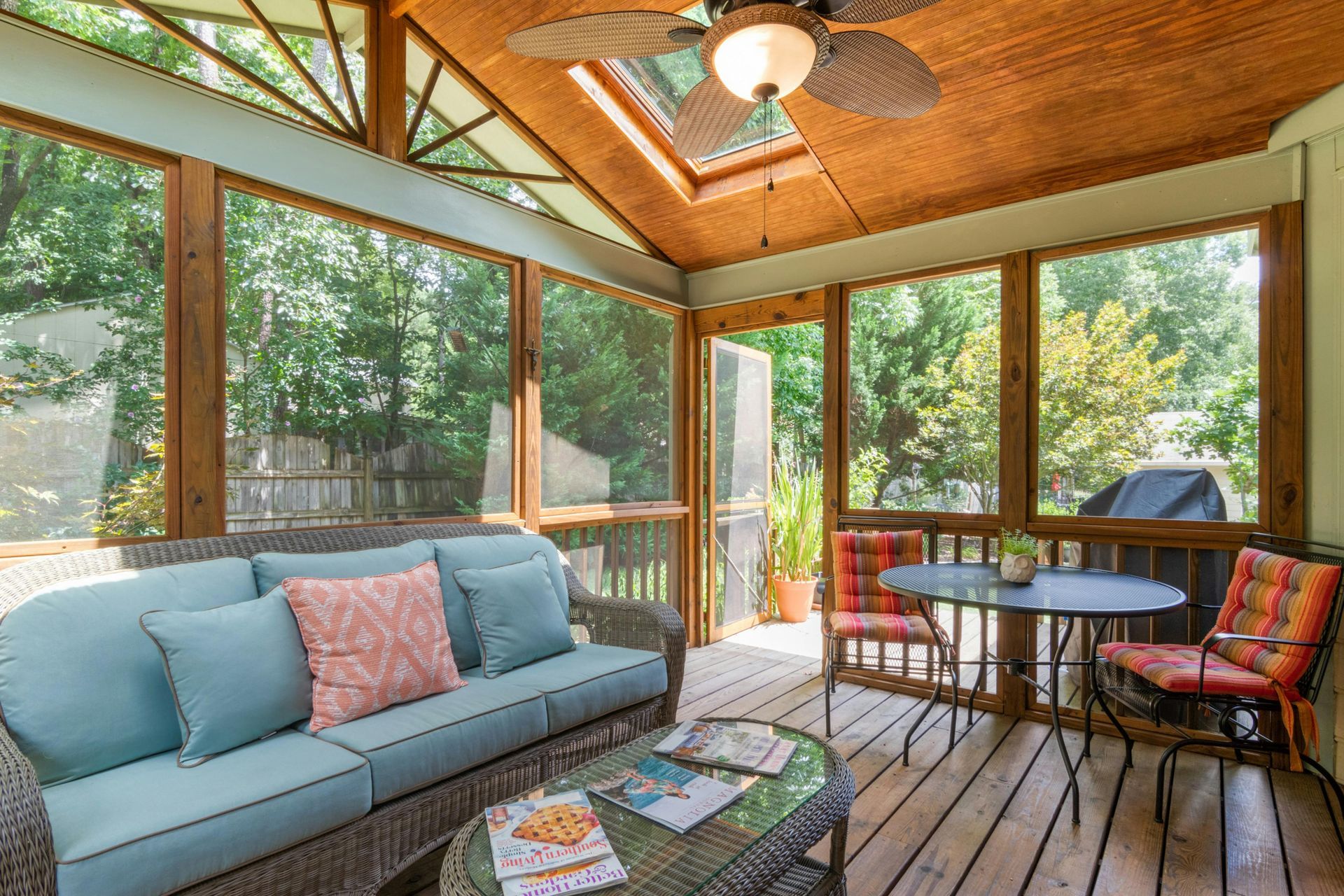 A cozy screened-in porch with a blue sofa, a small dining table with chairs, and a ceiling fan under a wooden roof.