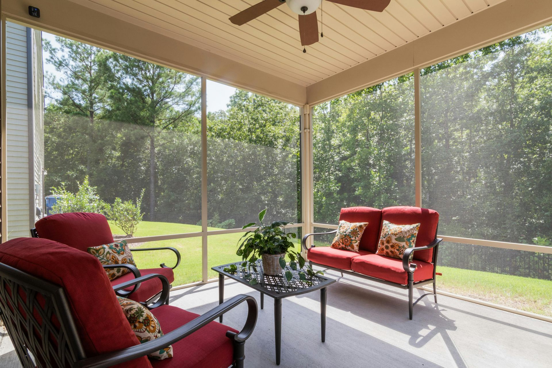 A screened-in porch with two red cushioned chairs and a small sofa, a center glass-topped table, and a ceiling fan.