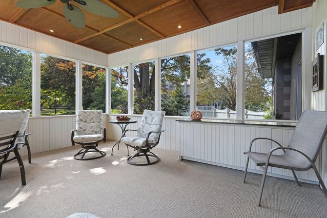 A bright, screened-in porch with light-colored walls, a wood-paneled ceiling, and several patio chairs around a small table.