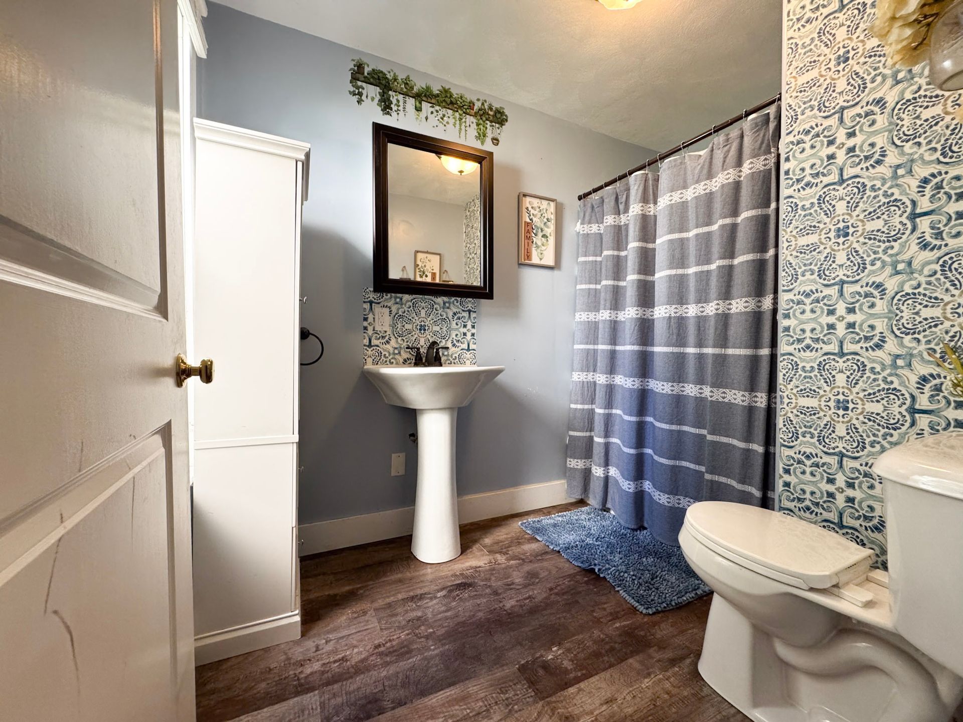 Small bathroom with white pedestal sink, toilet, shower curtain, mirror, and blue patterned wallpaper.