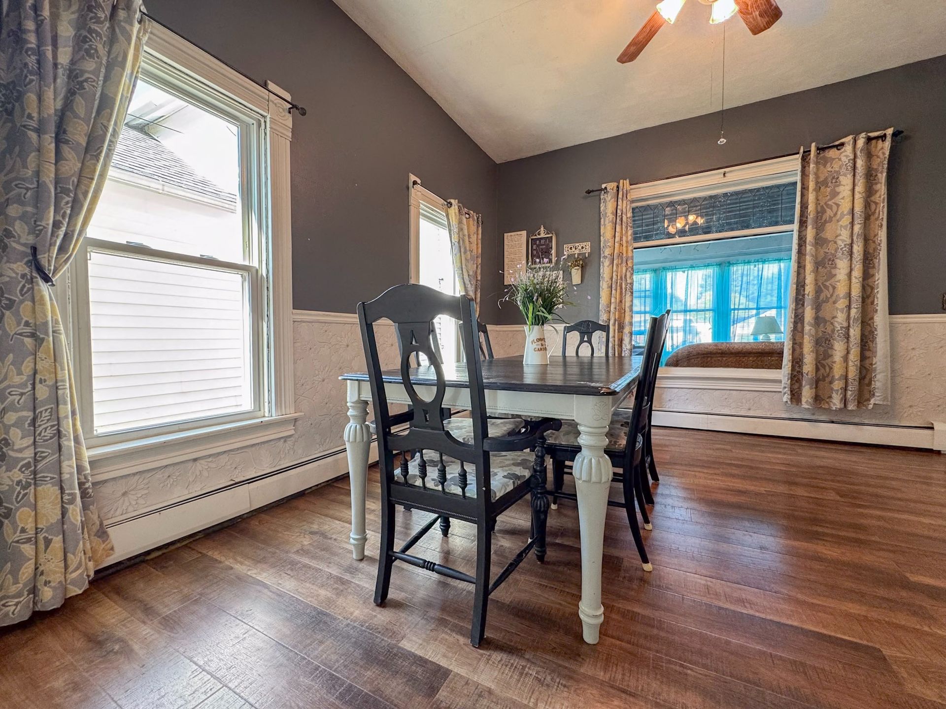Dining room with dark wood floor, white table, and chairs near windows with patterned curtains.