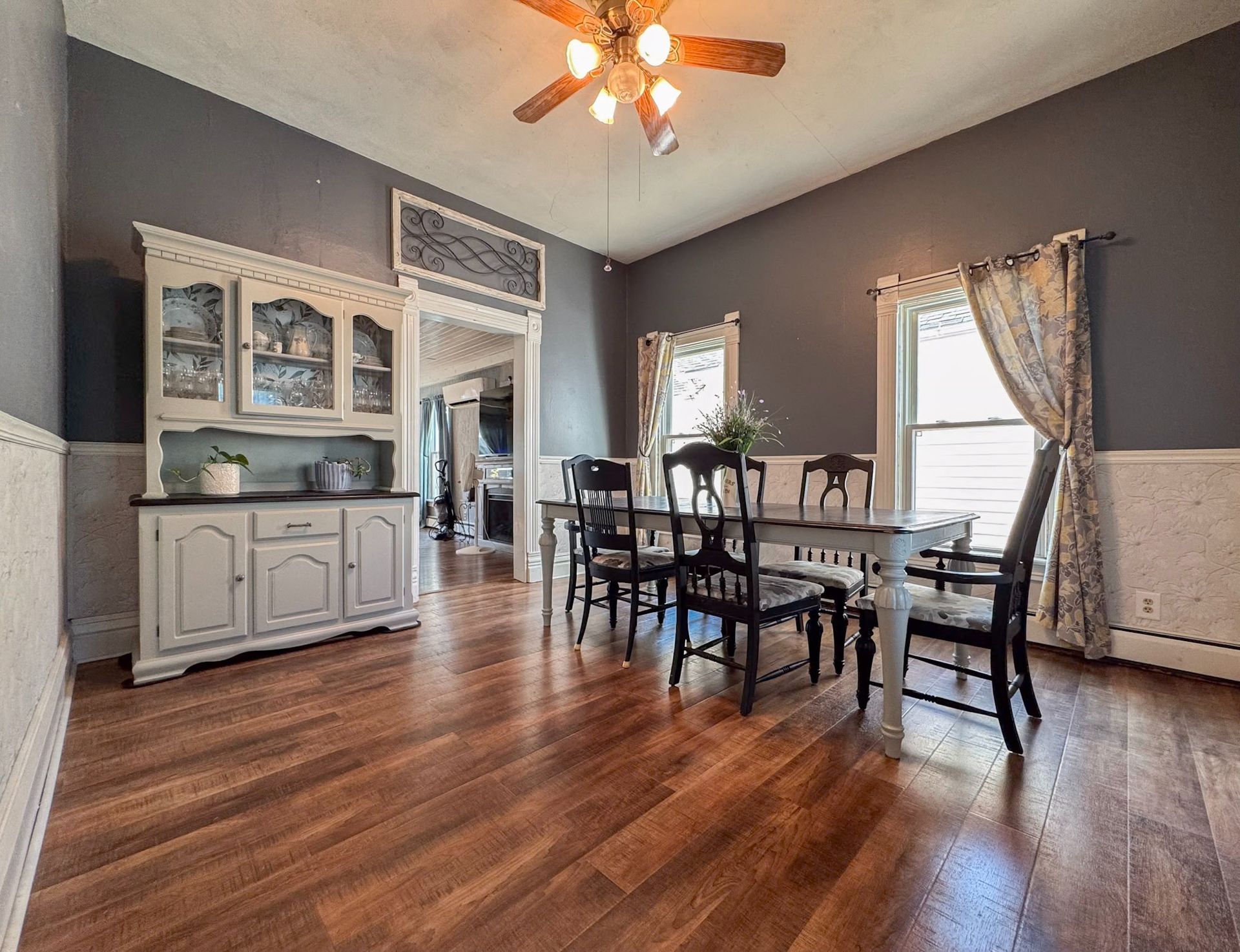 Dining room with dark wood table, gray walls, white cabinet, and large windows with curtains.