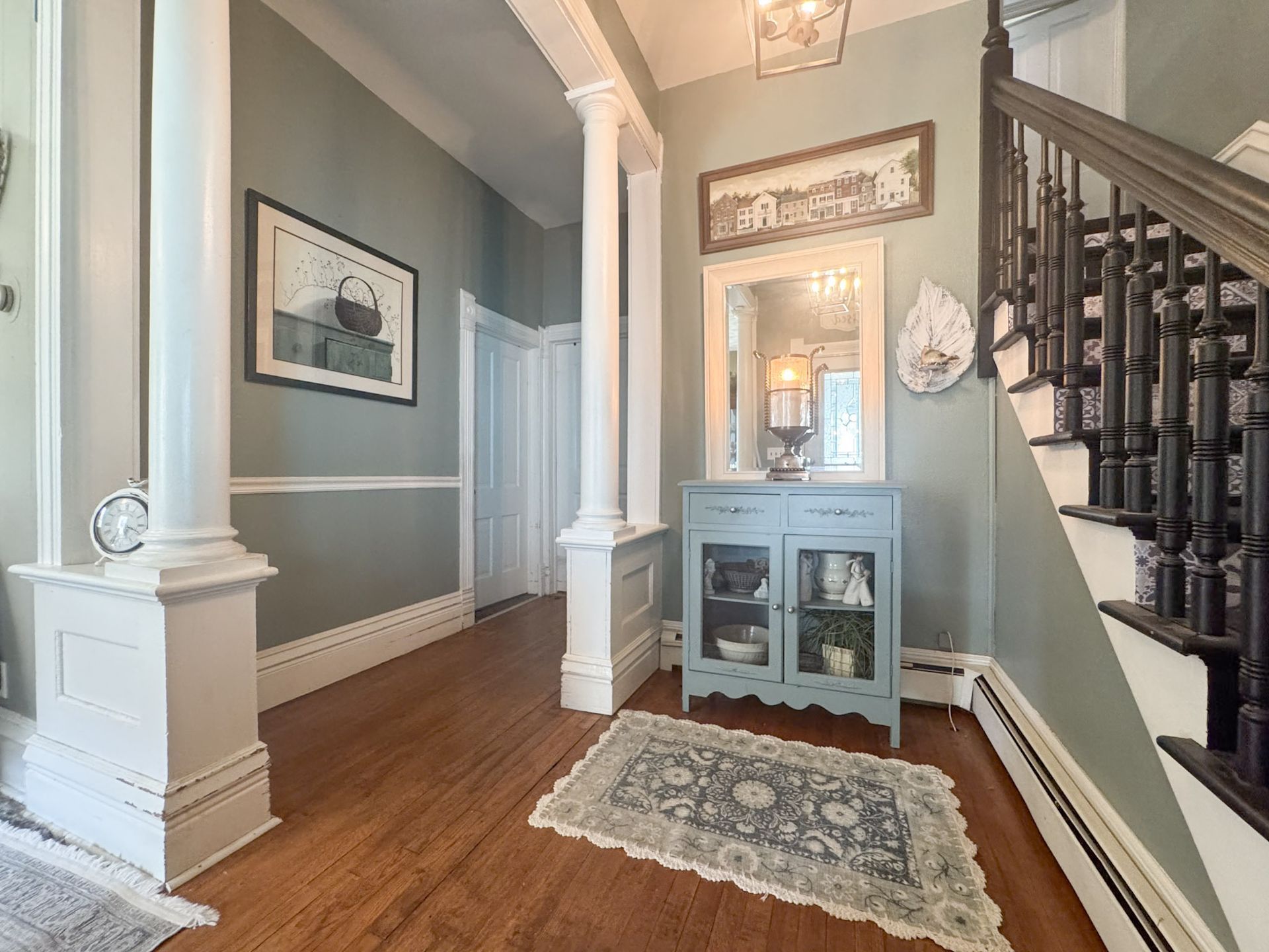 Bright entryway with hardwood floors, white columns, staircase, mirror cabinet, and framed wall art.