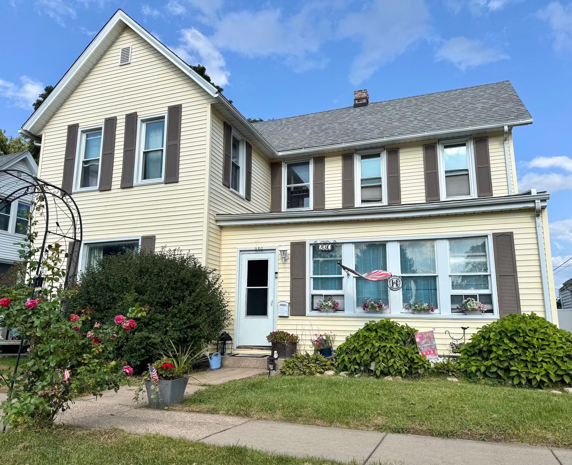 Suburban two-story beige house with blue shutters, white front door, and a flower garden in front.