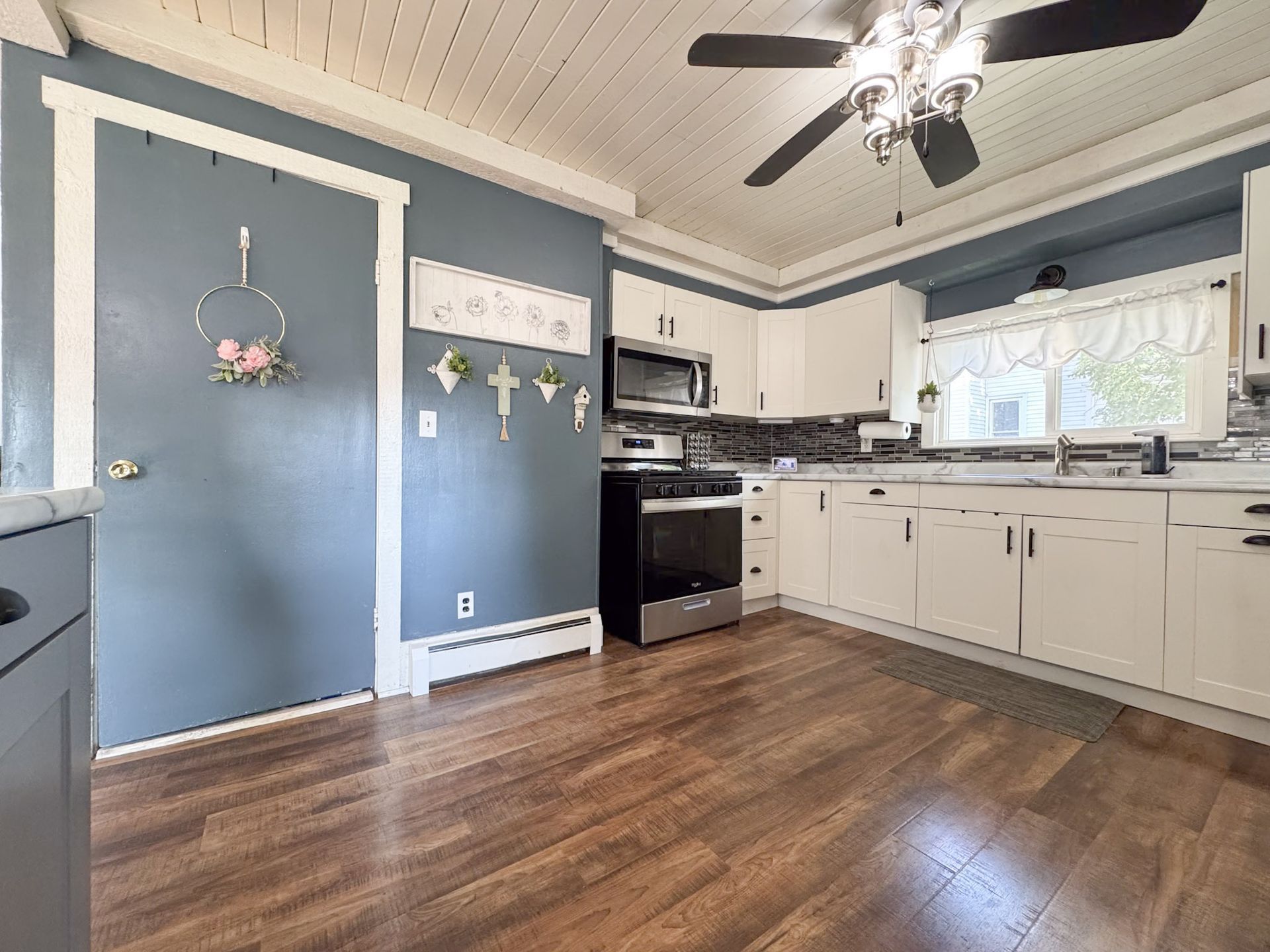 Blue-walled kitchen with white cabinets, dark wood floor, and ceiling fan in a bright interior.