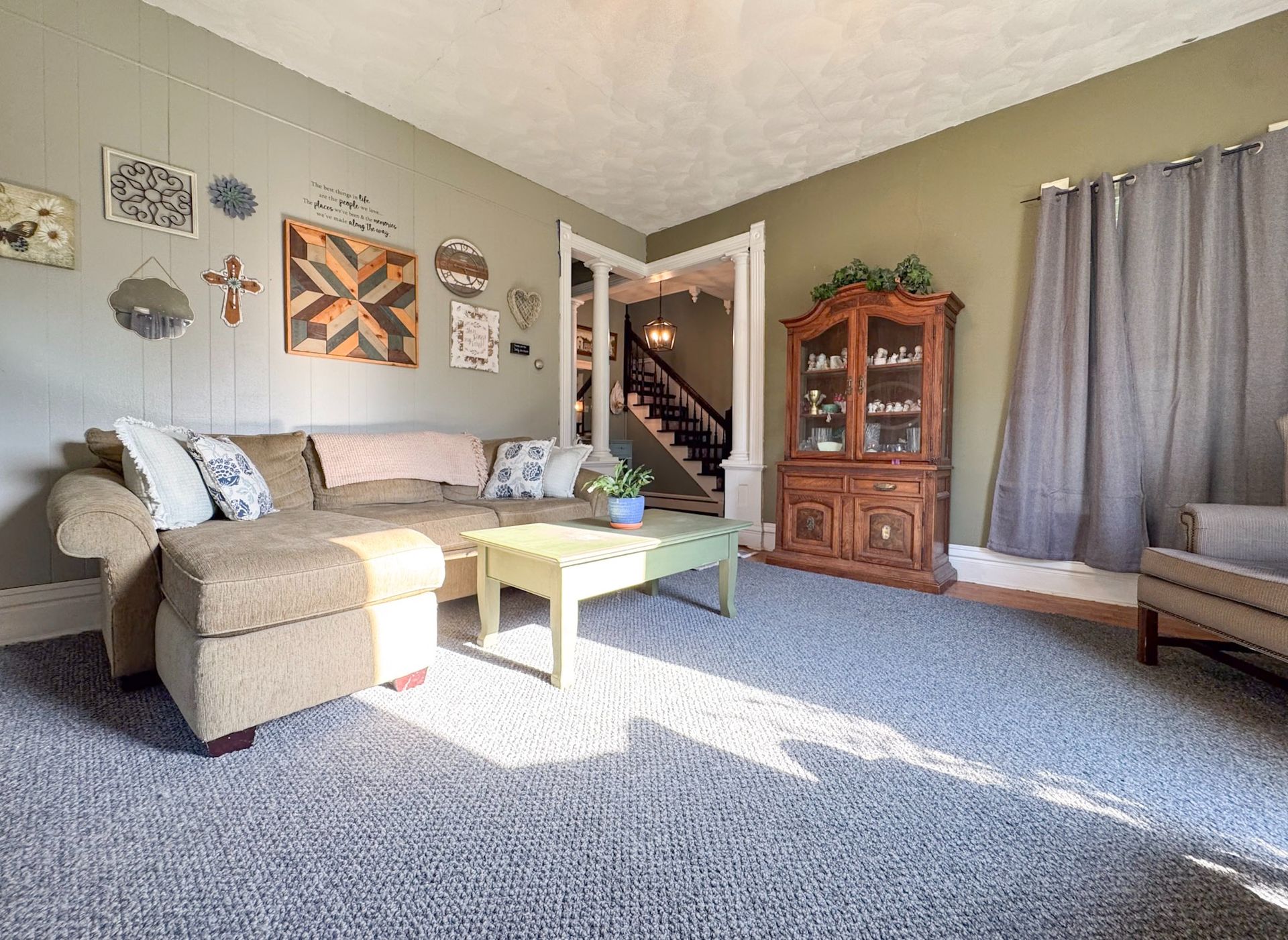 Bright living room with beige sectional, coffee table, cabinet, stairs, and sunlight through curtains