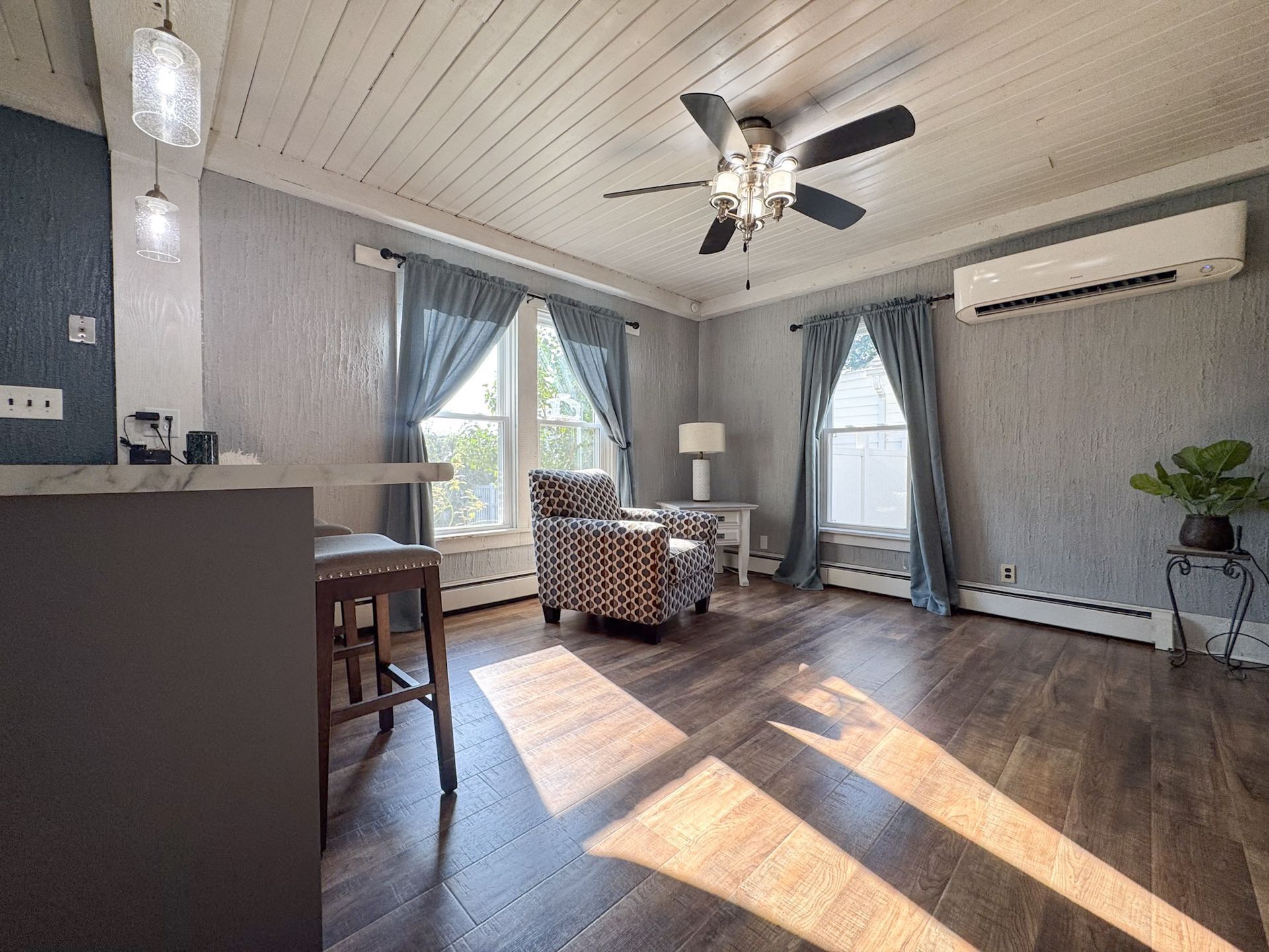 Bright living room with gray walls, large windows, wooden floor, and a ceiling fan casting sunlight.