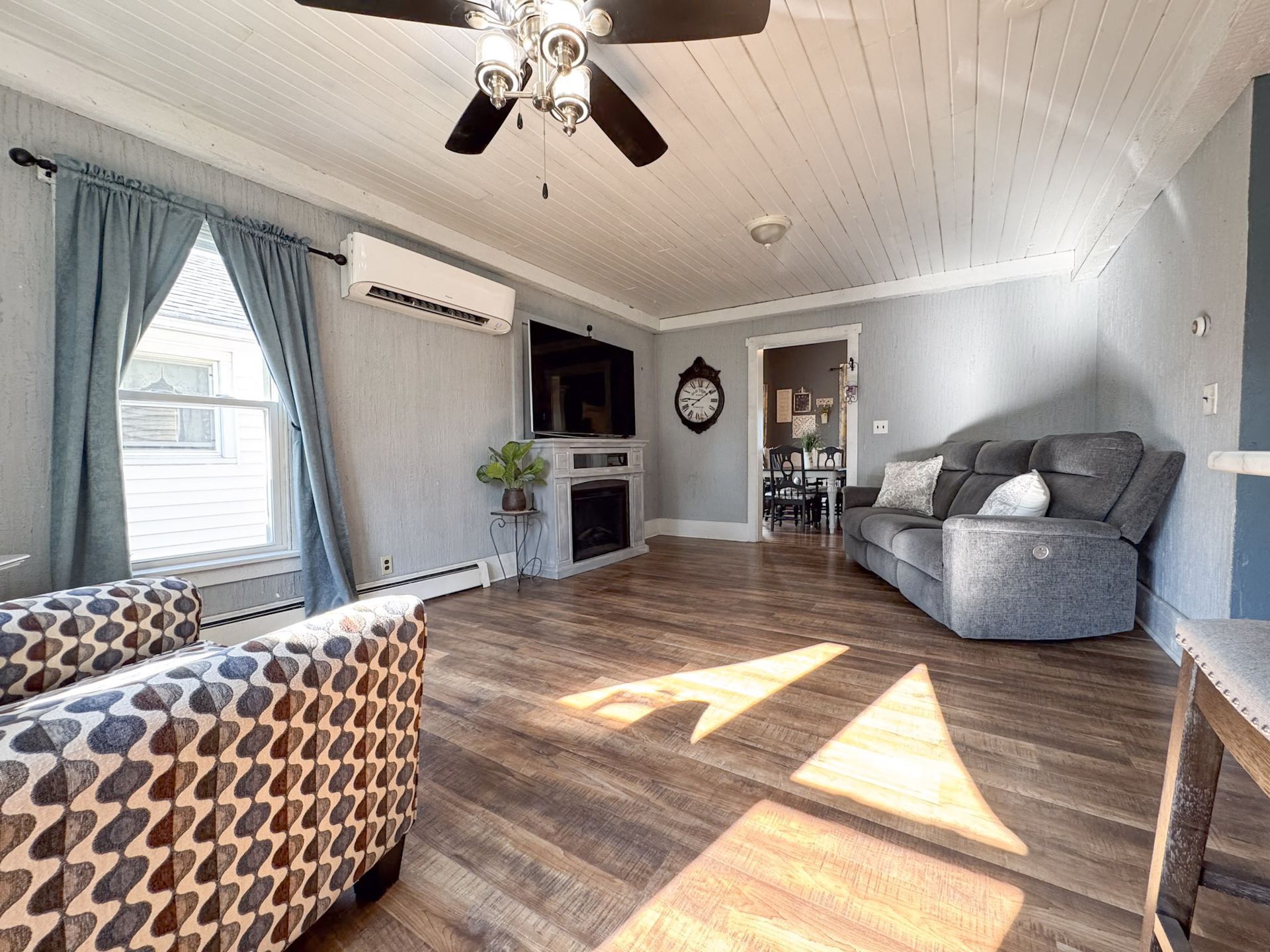 Bright living room with gray walls, wood floors, fireplace, gray sofa, patterned chair, and sunlight on the floor.