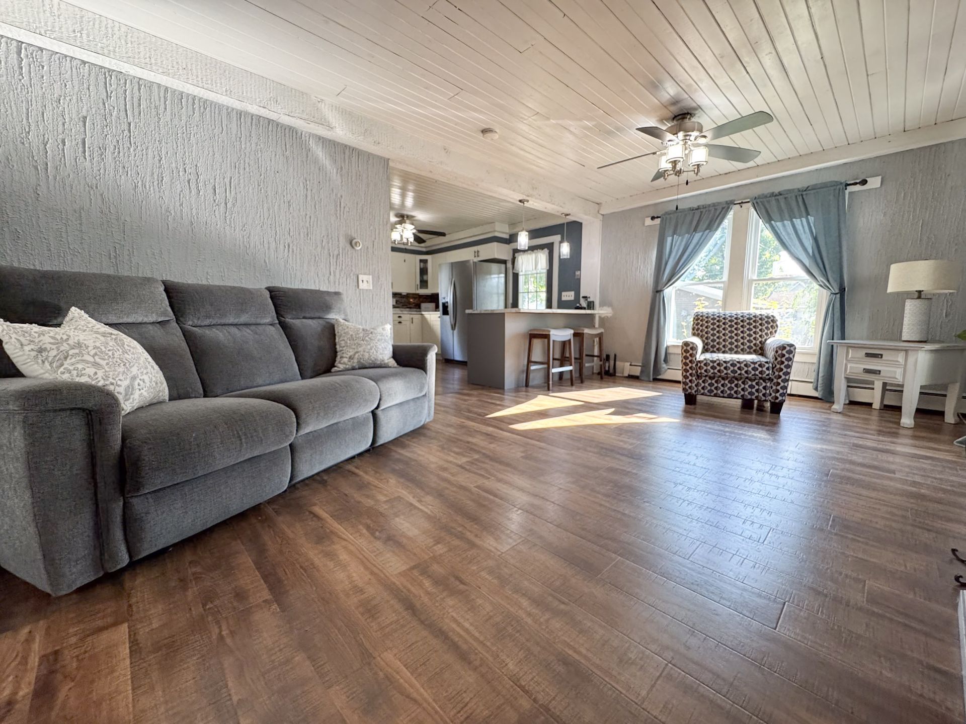 Bright living room with gray sofa, wood floors, fireplace, and large curtained window with armchair.