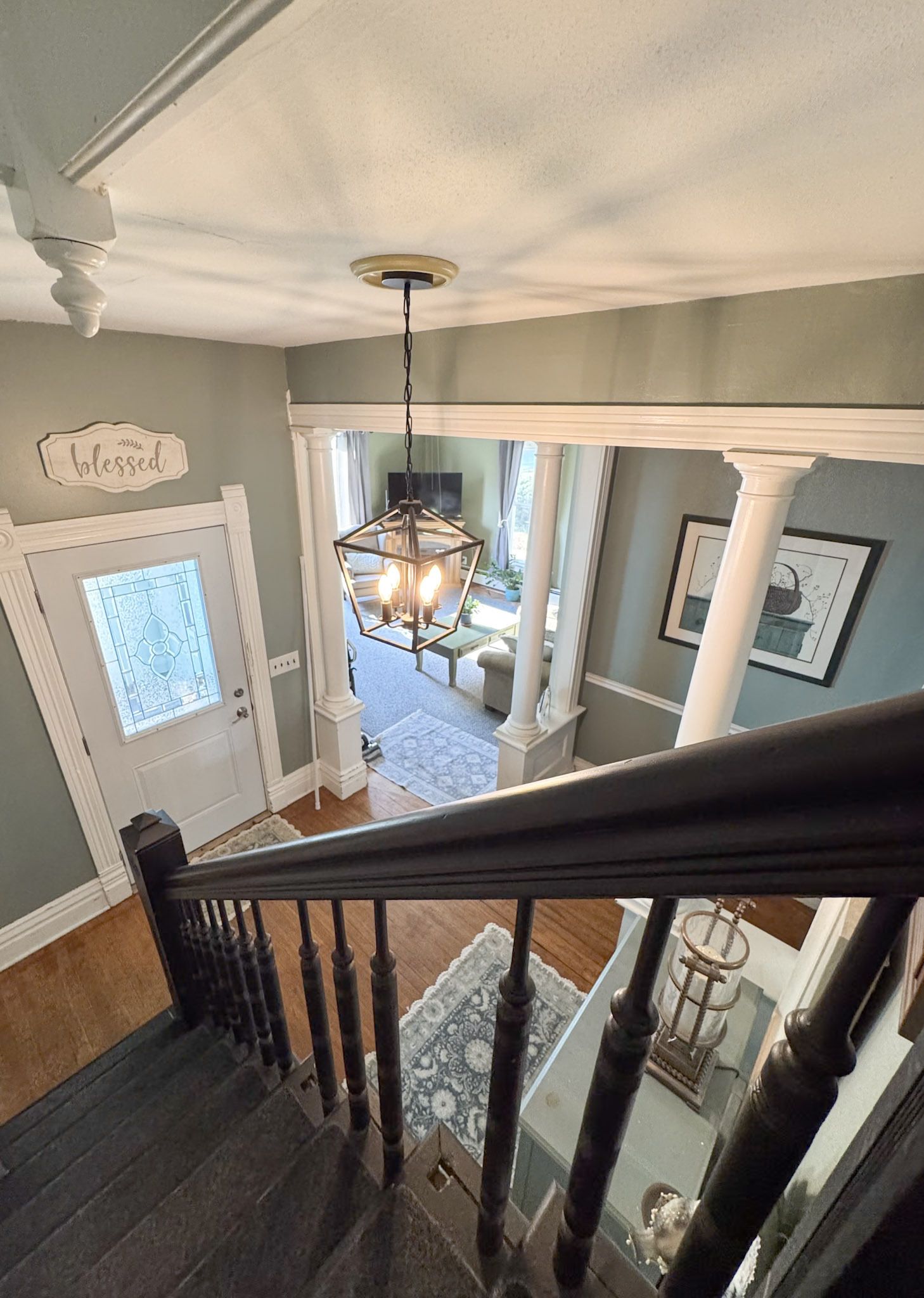 Staircase landing with dark wood railing, hanging light, and view into a bright foyer with gray walls.