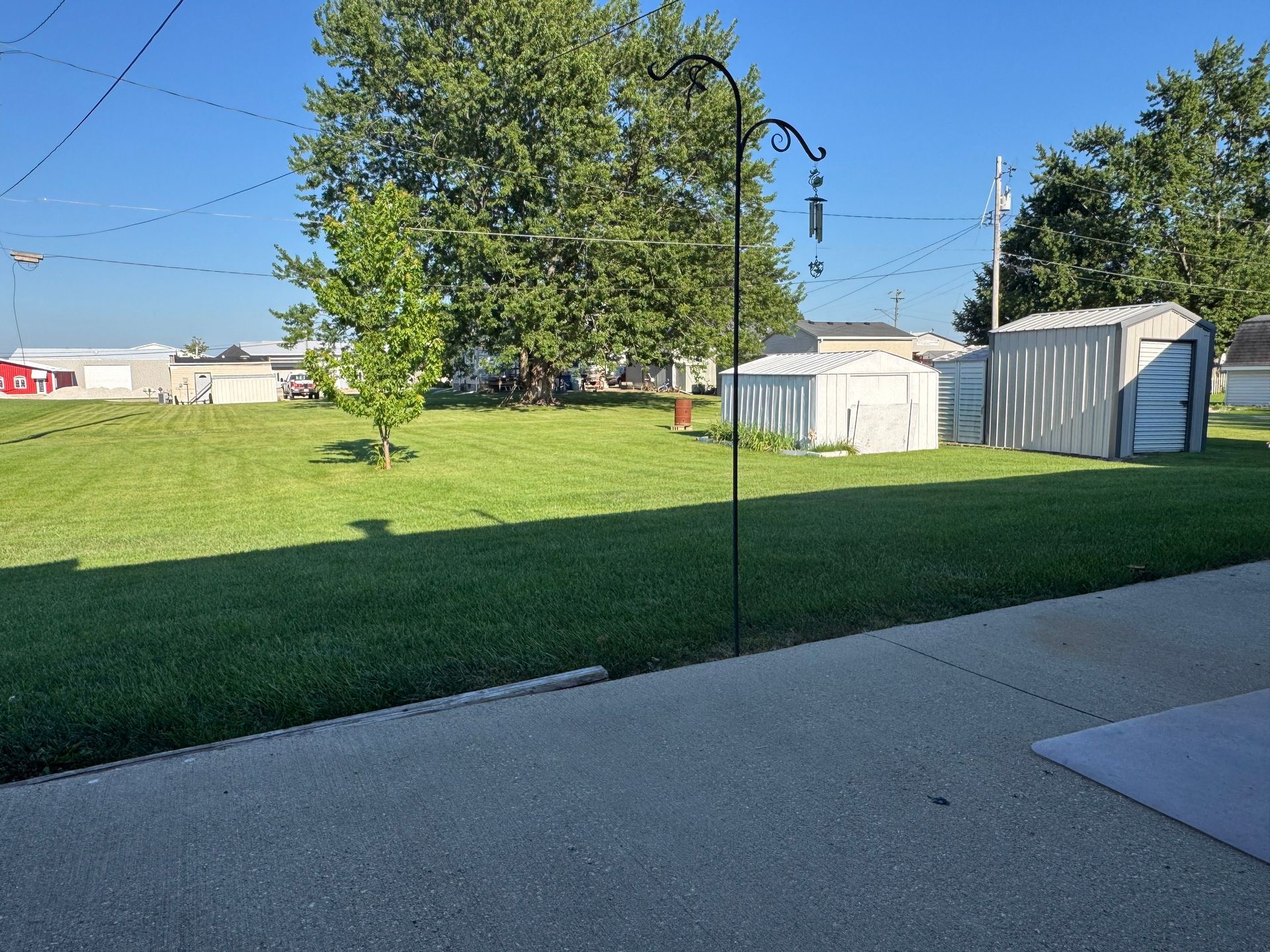 Backyard lawn with a bird feeder pole, shed, trees, and a concrete patio in bright sunlight