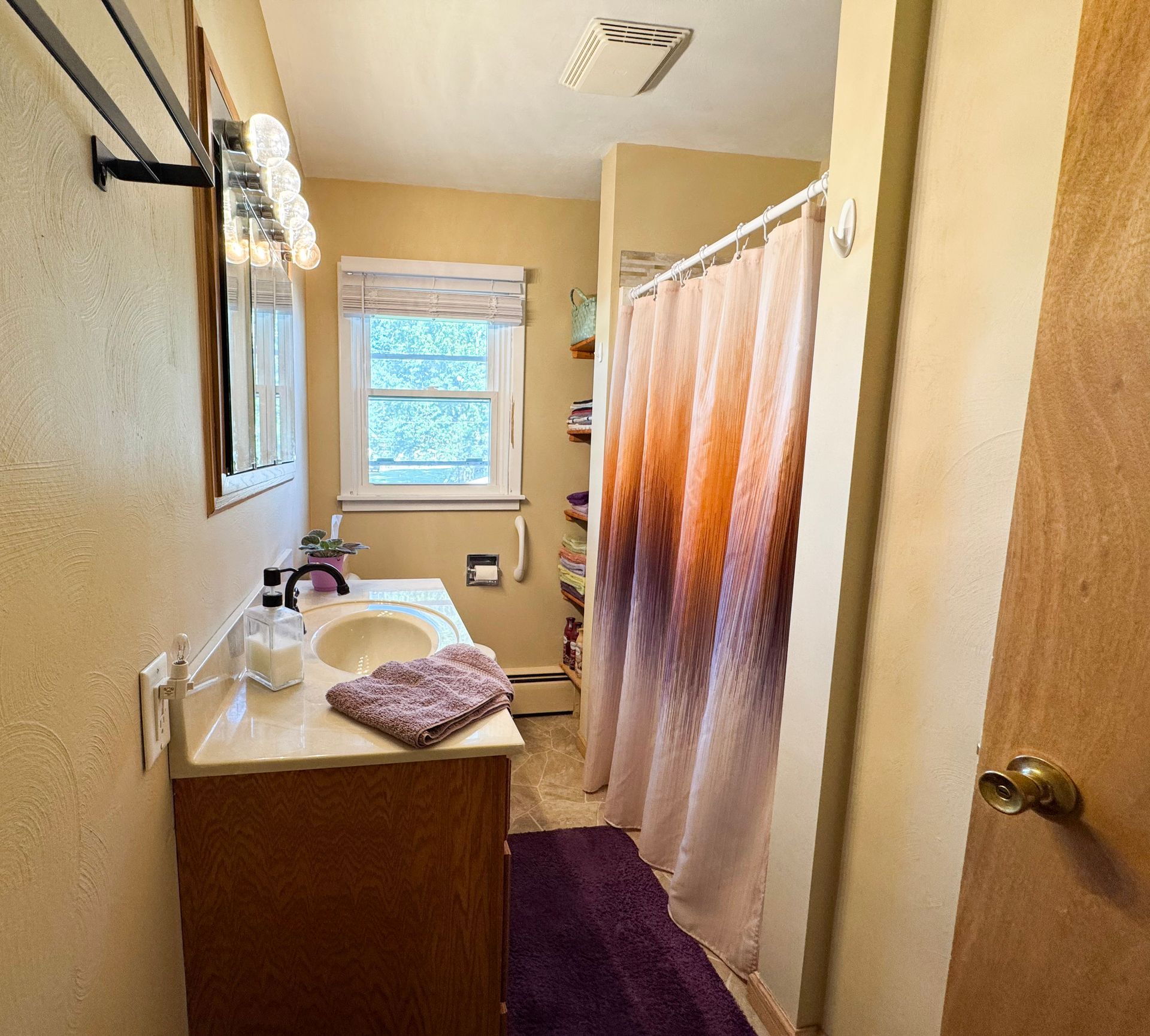 Small bathroom with sink, mirror, window, and peach shower curtain; purple rug and towels.