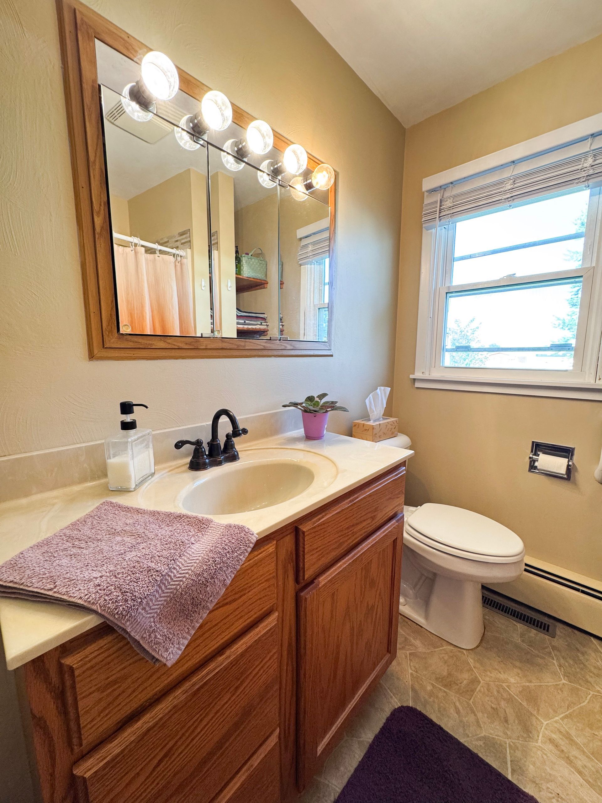 Small beige bathroom with wooden vanity, mirror lights, sink, toilet, and window blinds.