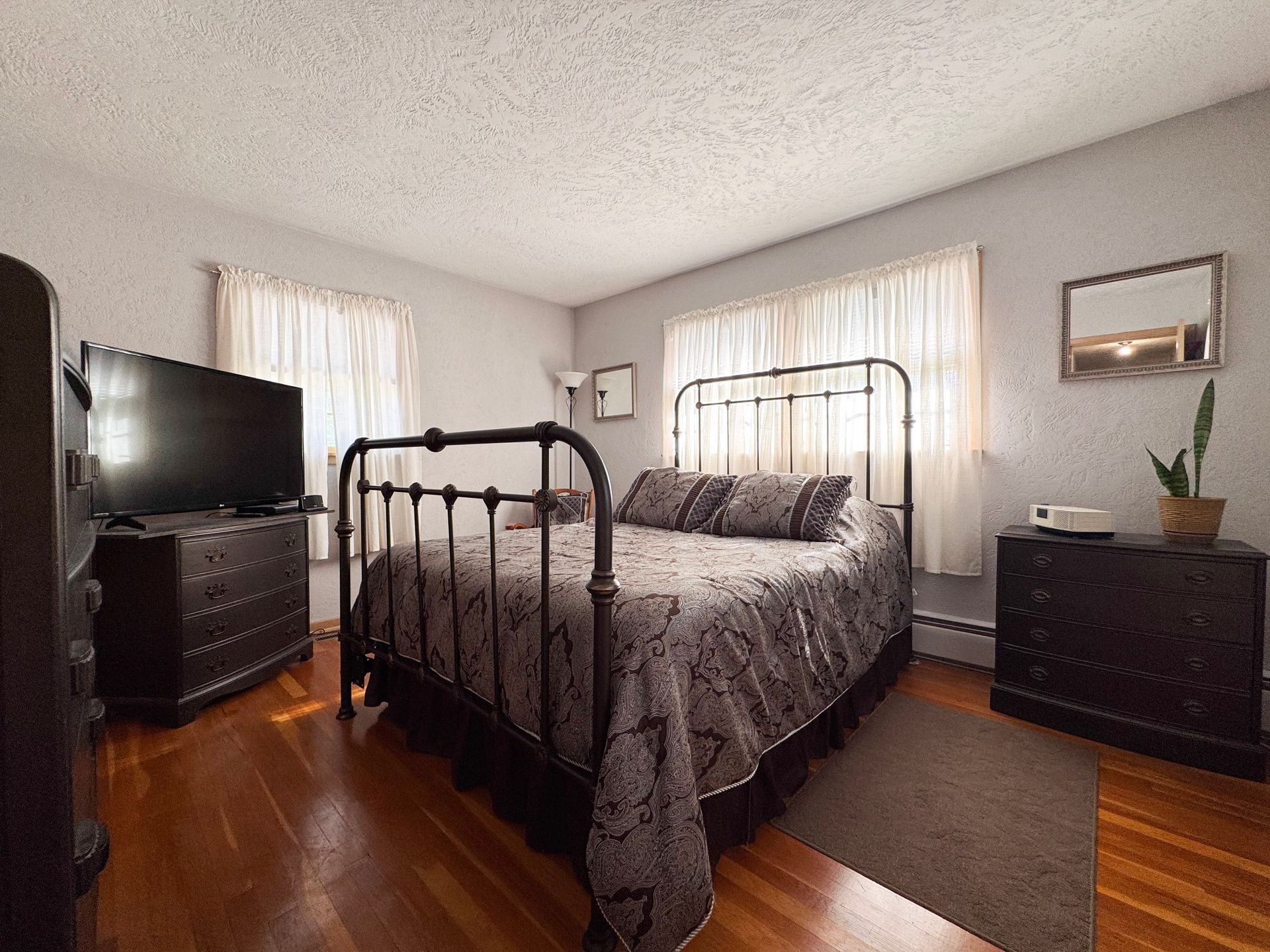 Bedroom with black metal bed frame, gray bedding, hardwood floors, and two dressers by bright windows