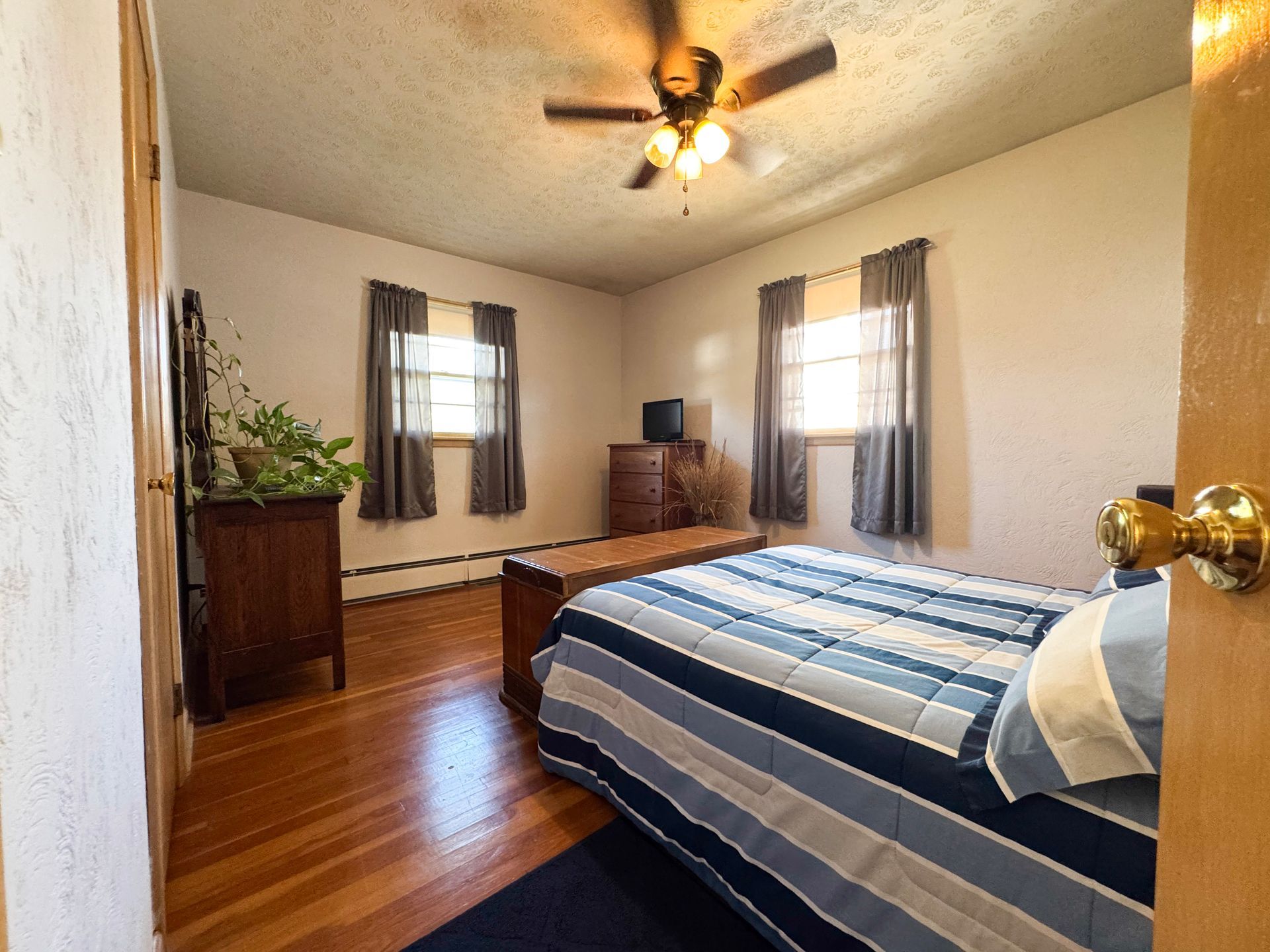 Bedroom with blue striped bedspread, hardwood floor, two windows, and a ceiling fan lights the room.