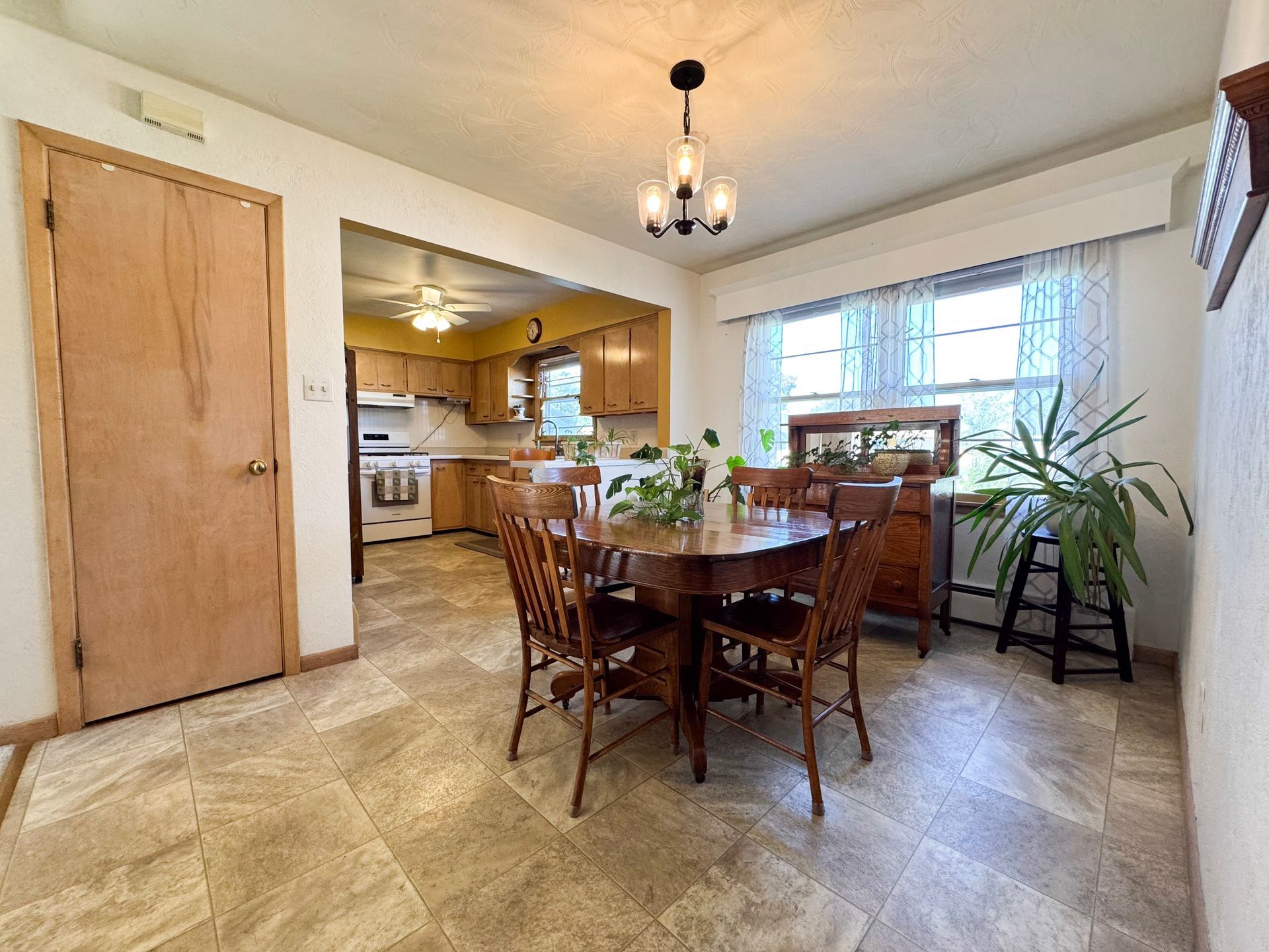 Dining room with wooden table and chairs, kitchen visible, large window, and a potted plant.