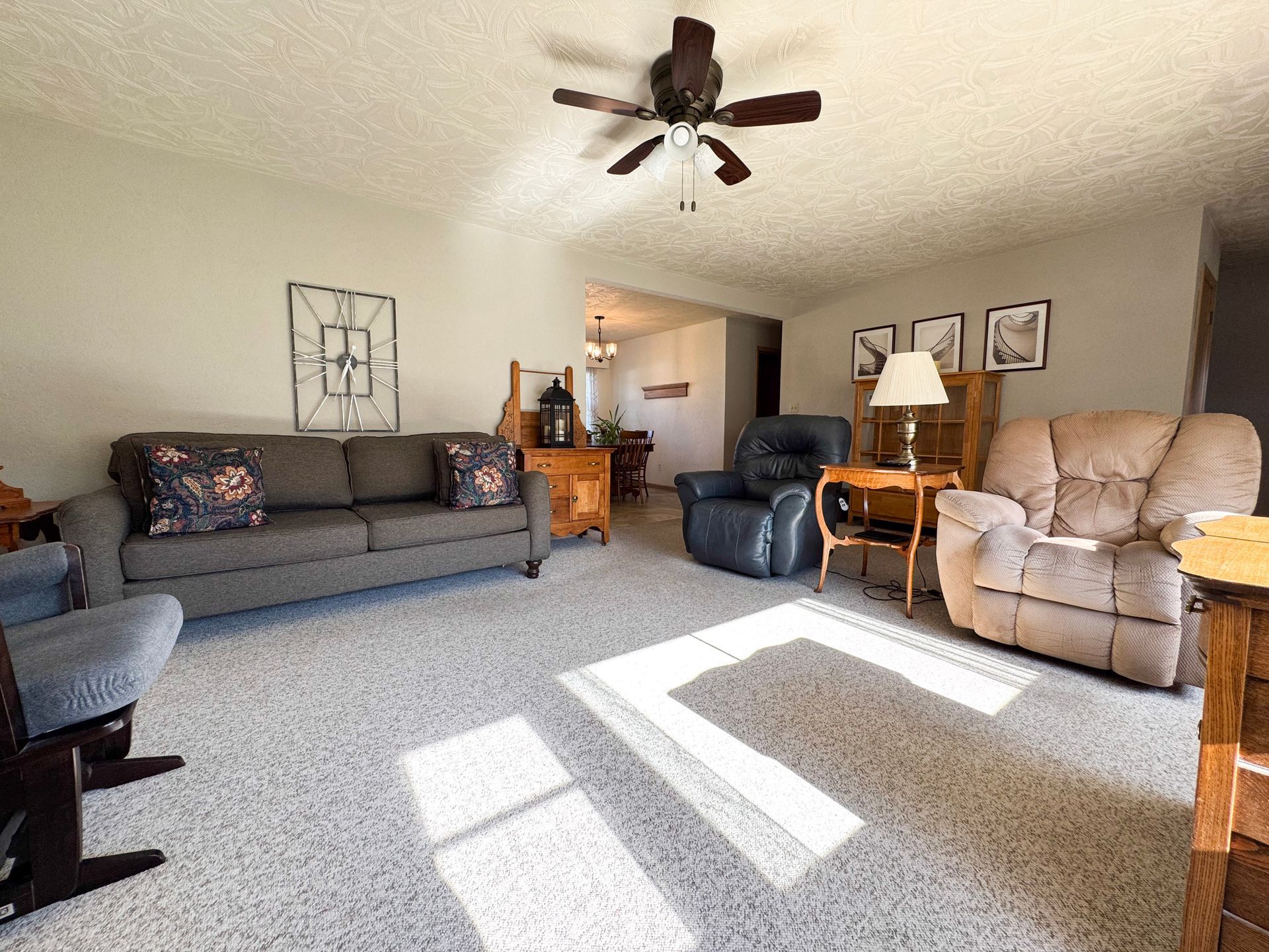 Bright living room with gray sofa, blue recliner, beige armchair, ceiling fan, and sunlight on carpet.