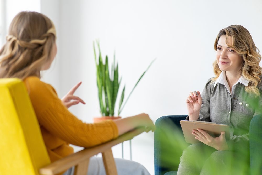 A woman is sitting in a chair talking to another woman while holding a tablet.