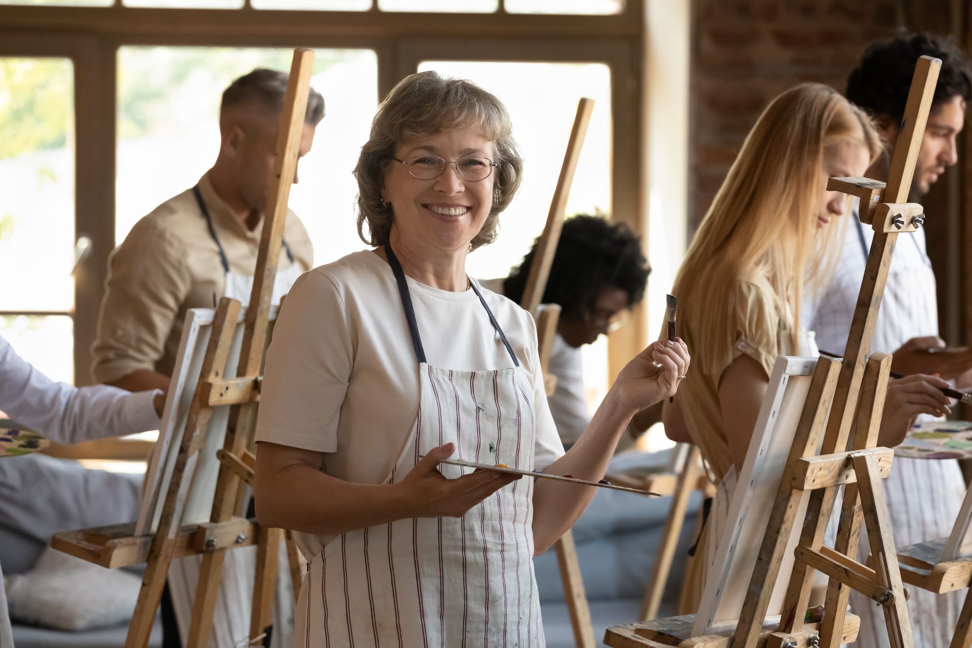 An eldery white woman takes a painting class, her younger classmates can be seen behind her in the studio, she asks herself the question