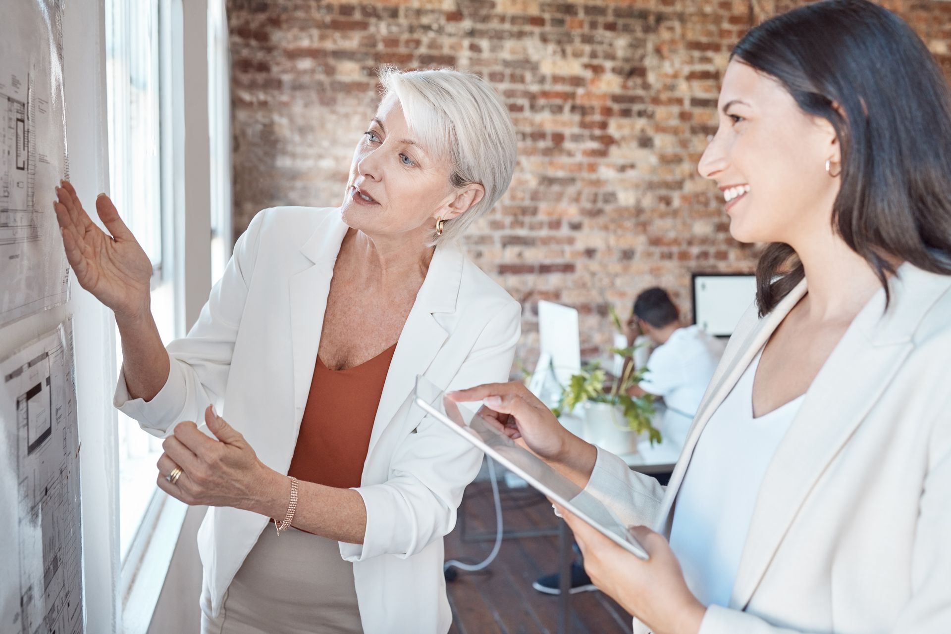 Two women are looking at a whiteboard in an office working on a project and practicing communication in leadership