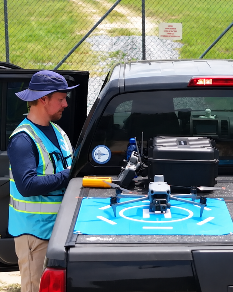 Worker in blue safety vest inspecting equipment on the back of a black truck near a fenced area.