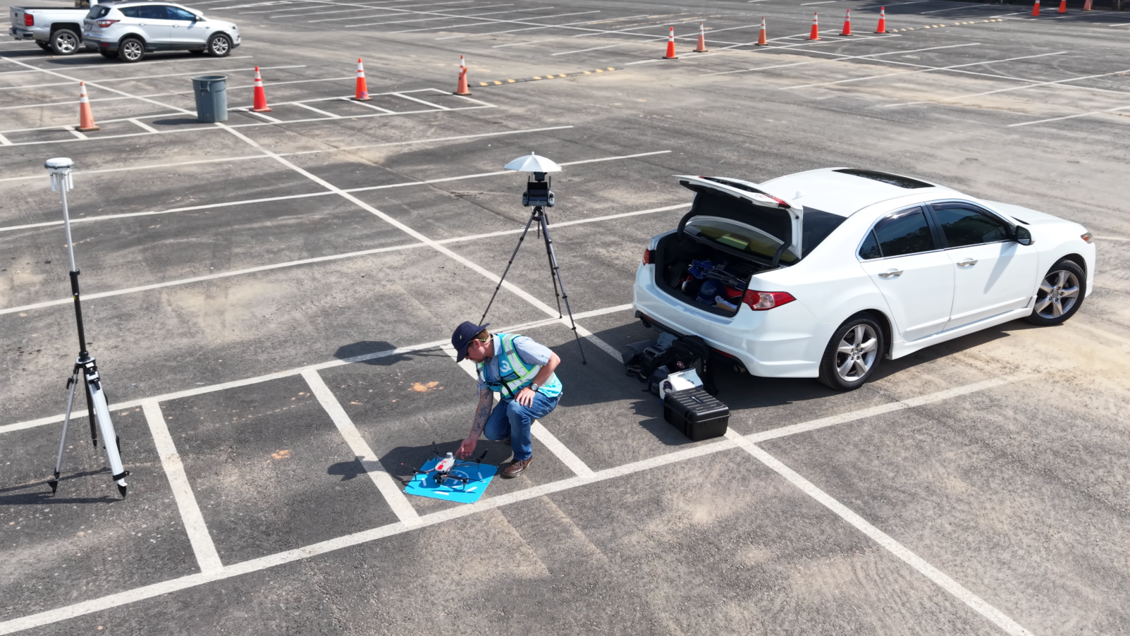 Person working with equipment beside a white car in a marked parking lot, with cones and tripods nearby.