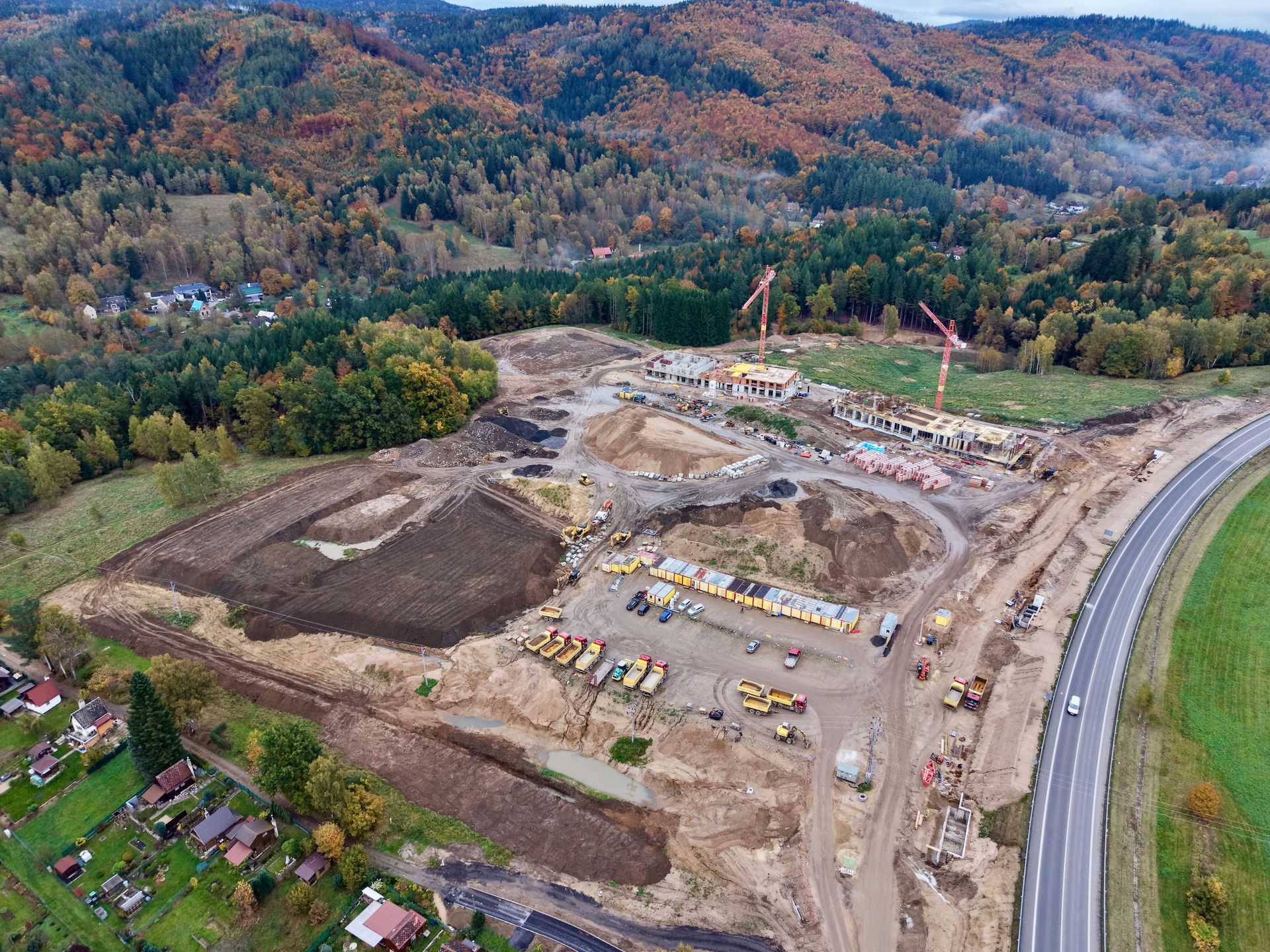 Aerial view of a large construction site beside a road, with heavy equipment and forested hills beyond.