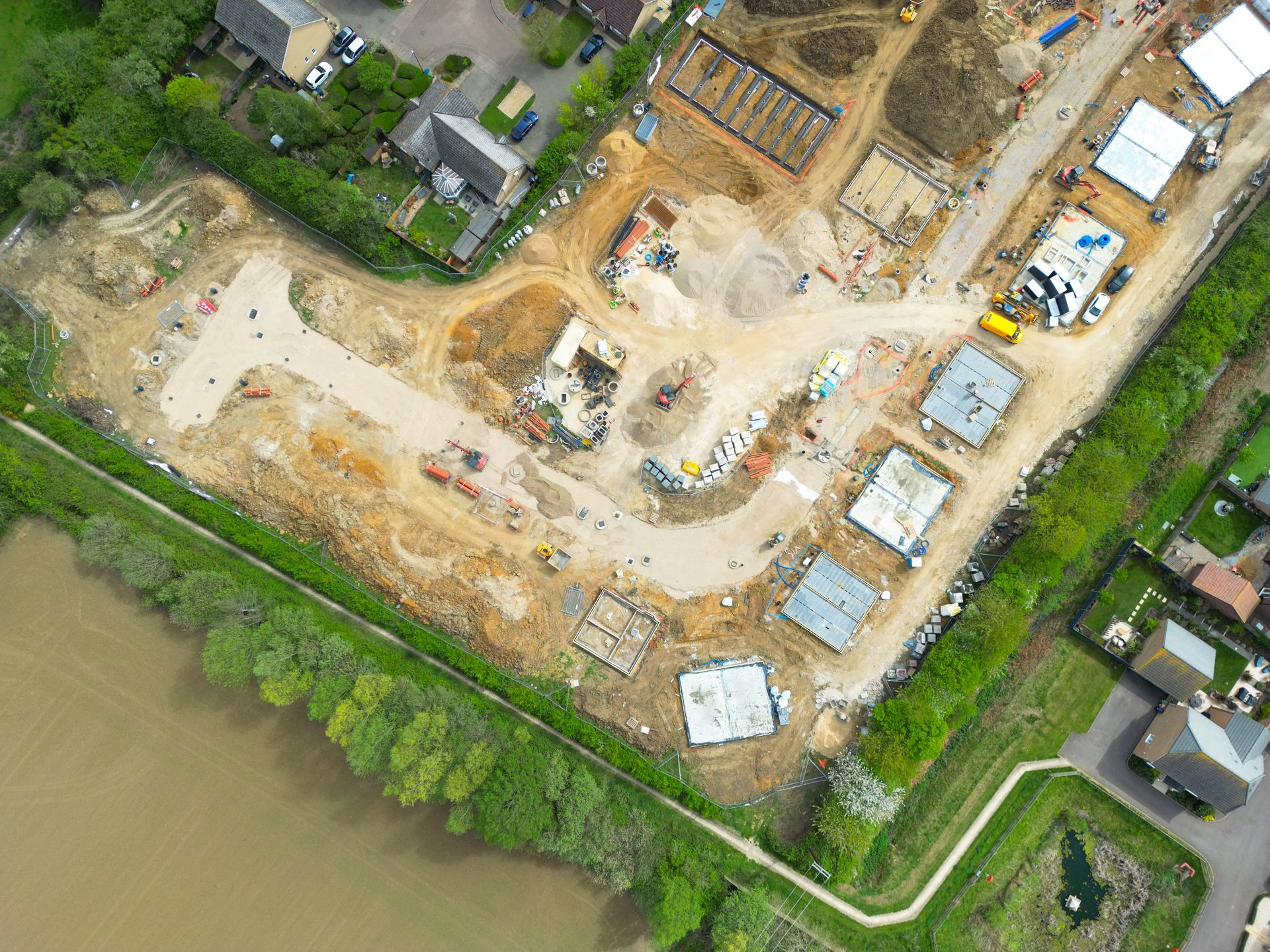 Aerial view of a construction site beside a river, with dirt lots, building foundations, and nearby houses.