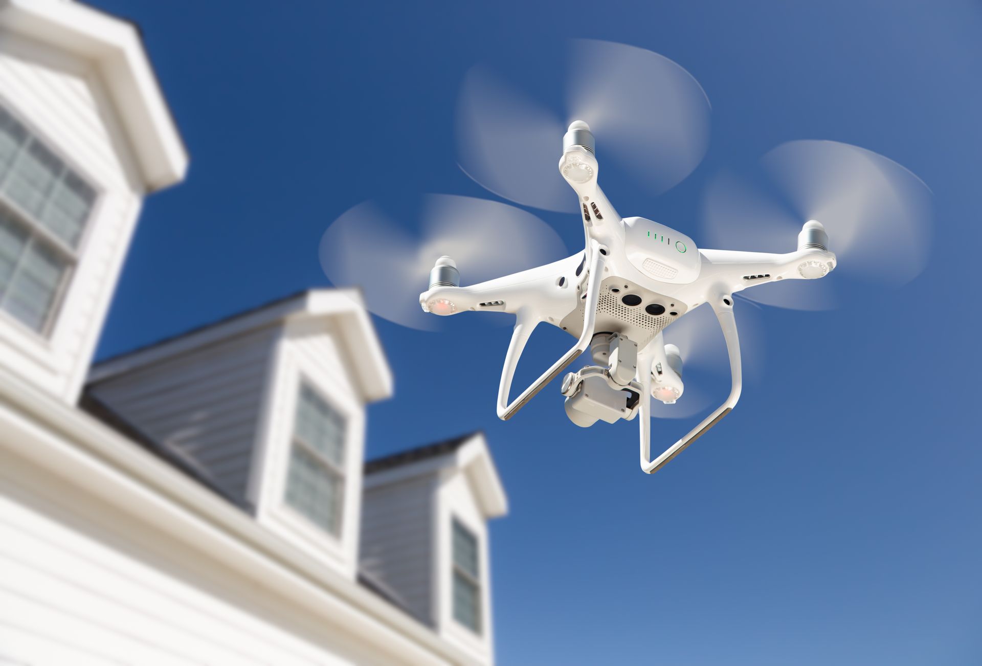 White quadcopter drone flying above a house roof against a blue sky