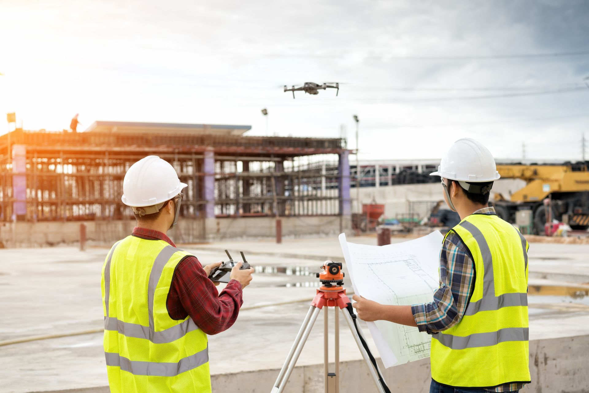 Two construction workers in hard hats reviewing plans at a building site with a drone overhead