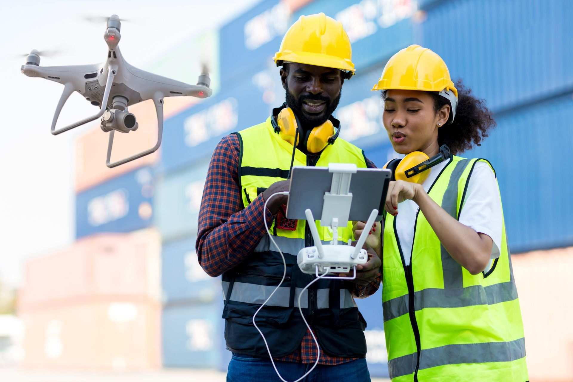 Two workers in yellow safety vests and helmets operating a drone controller at a shipping yard