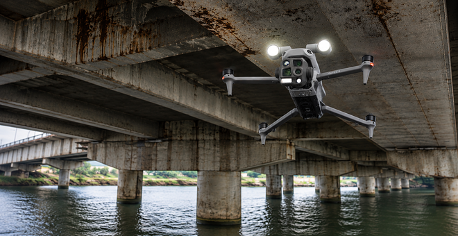 Drone hovering under a bridge over water, with concrete pillars and city structures in the background