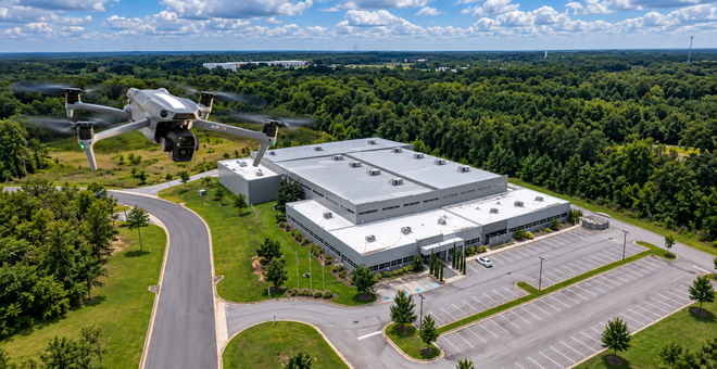 Drone flying over a white industrial building and parking lot beside a winding road, surrounded by dense trees