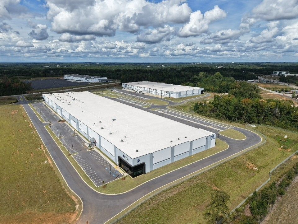 Aerial view of a large white warehouse in a grassy industrial area under a cloudy sky.