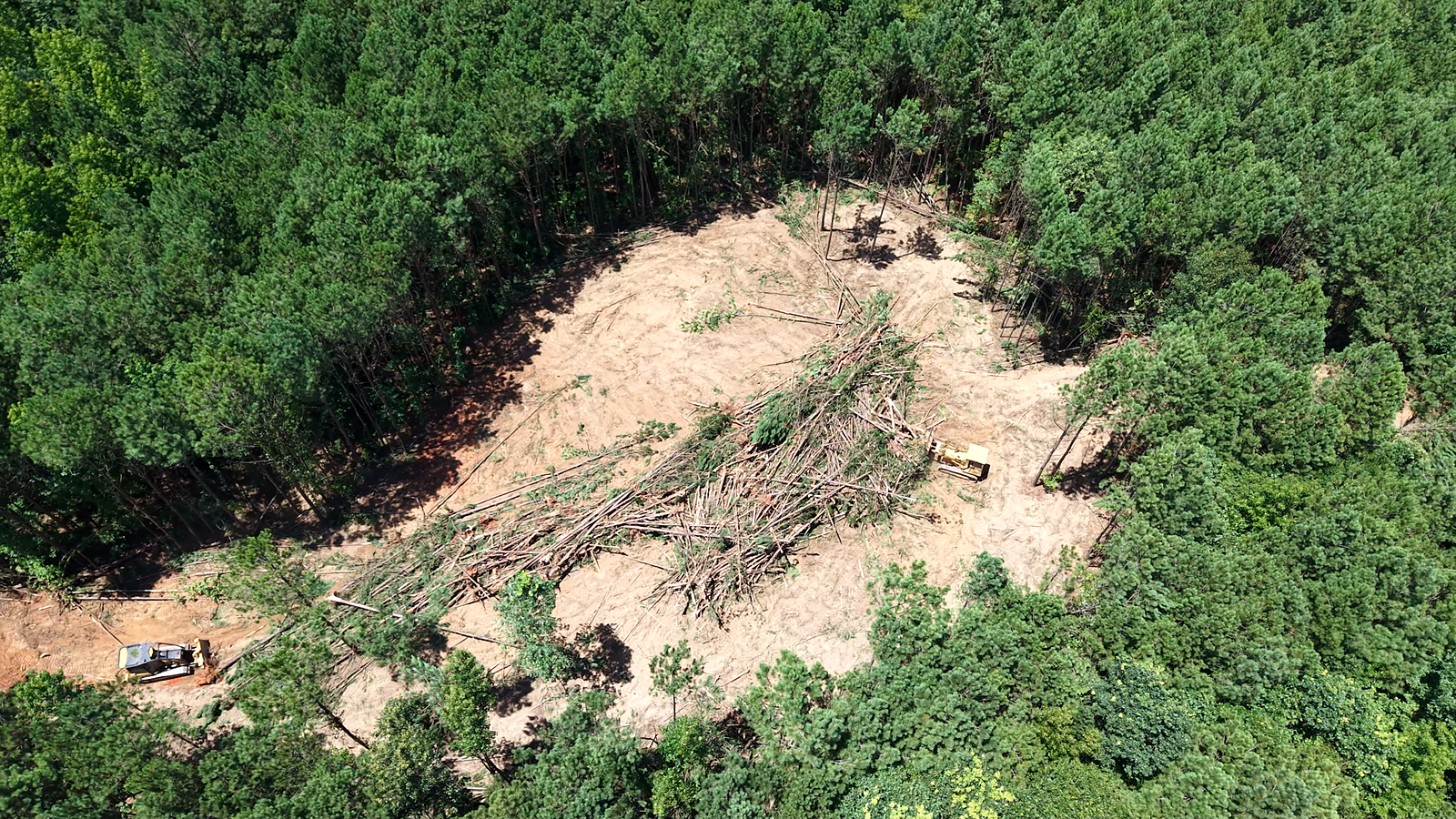 Aerial view of a large tan clearing in dense green forest with debris and a few vehicles nearby
