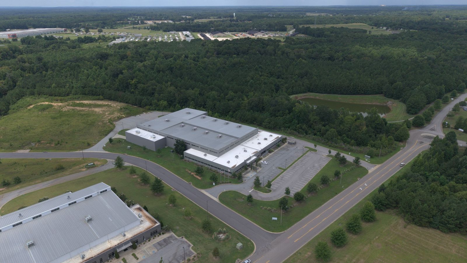 Aerial view of a large white industrial building beside a road, surrounded by trees and parking lots.